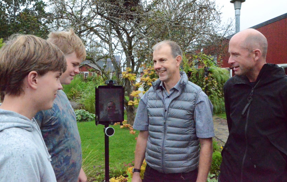 Flera personer står runt en robot som är en slags segway med en skärm på. På skärmen syns en femte person som kan vara närvarande i undervisningen även fast hen är hemma.