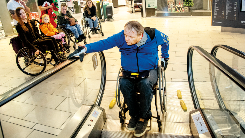 En person sitter i en rullstol och ska åka upp för ett rullband.