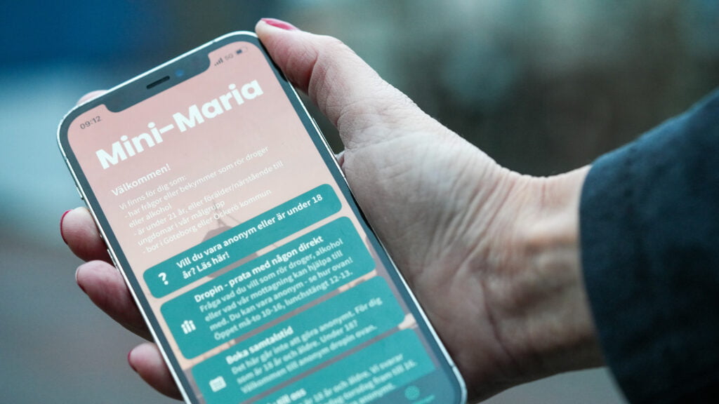 En hand håller i en mobil med appen Mini-Maria online