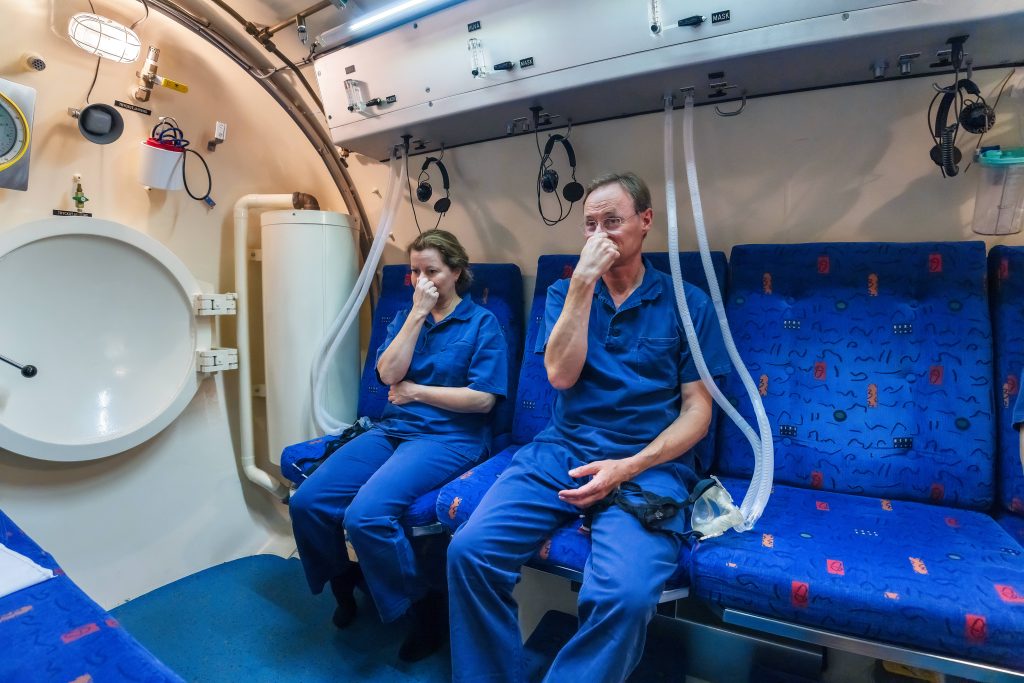 Två personer sitter i en tryckkammare. De håller för sina näsor så att de kan tryckutjämna.